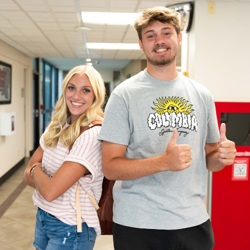 Two students giving the thumbs up in a hallway on campus.